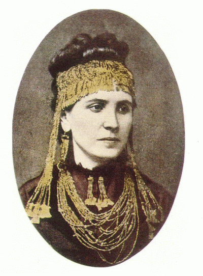 Portrait photo of Schliemann, a young woman, wearing an elaborate draping gold headdress, large gold earrings, and a loopy large gold necklace.