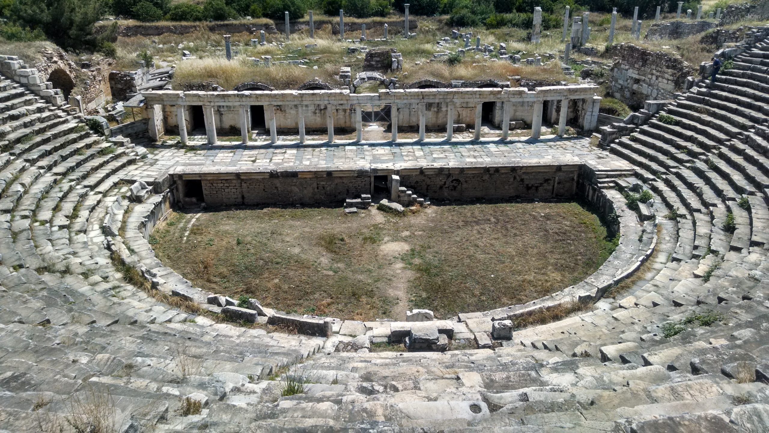 Stone amphitheatre, image taken from upper seats. Curved rows of stone benches descend to a semi-circular grass orchestra, backed by a raised stone stage. The lower story of an originally three story scaenae is preserved. It is aedicular style, with 14 marble columns. Behind the scaenae the ruins of other columns and buildings at the site can be seen.