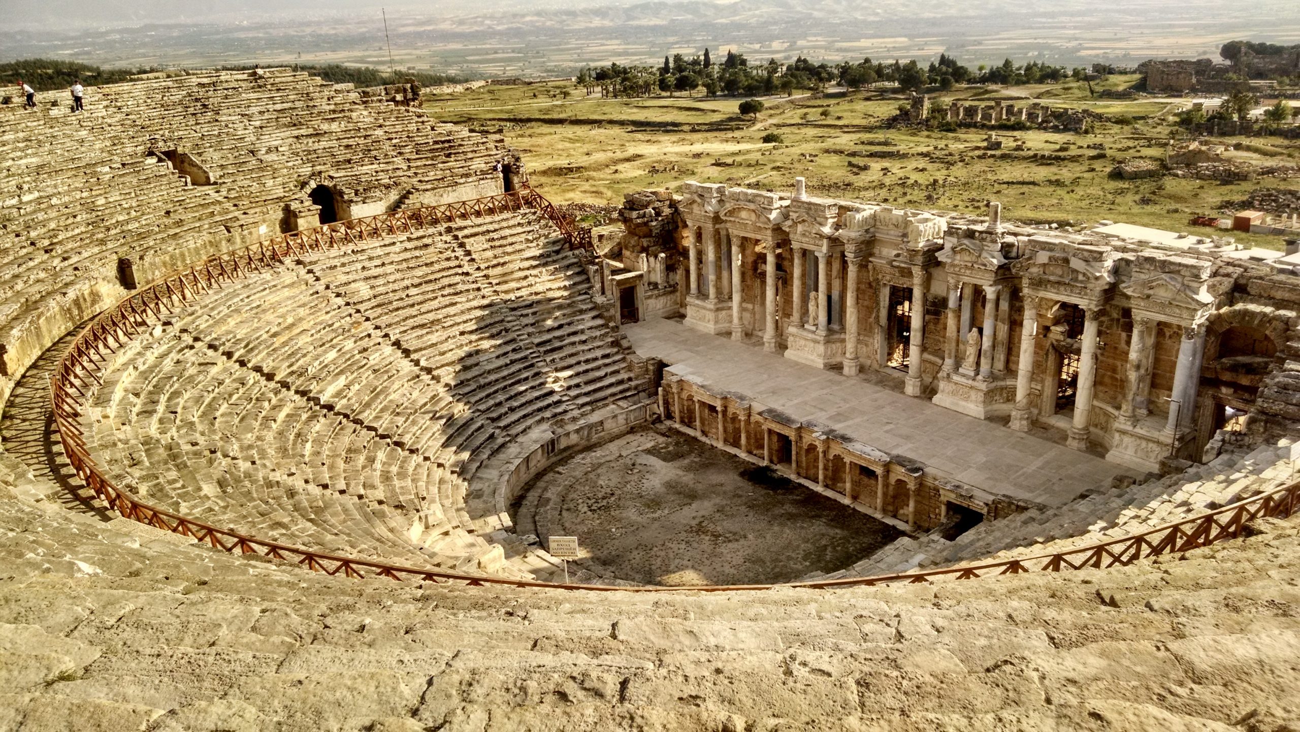 The theatre at Hierapolis in Pamukkale, Denizli, Turkey, picture taken from upper seats. Amphitheatre style, well-preserved stone theatre. Two large sections of semi-circular stone benches (45 rows in total) descending down a steep slope to a semi-circular orchestra at ground level. Ornate, small stone columns and niches in front of a raised, rectangular stone stage. The scaenae frons behind the stage is made up of ten ornate Corinthian columns, topped with entablature and forming six statuary niches with larger than life sized human figures in them. The surrounding country side behind the skene is flat, with greenish-brown dried grass and a copse of dark green trees off in the distance.
