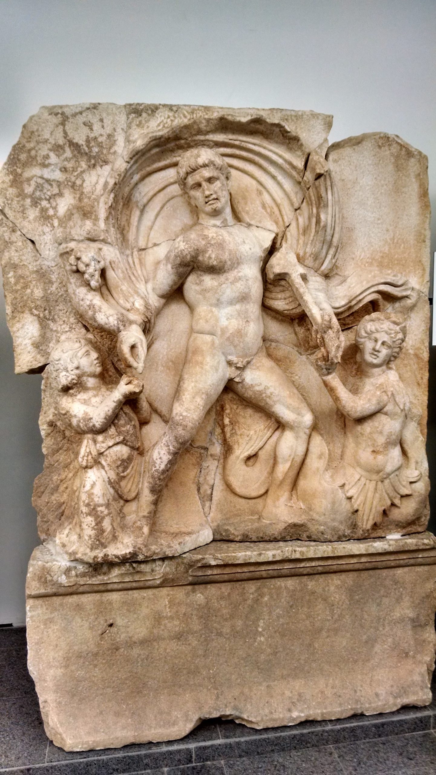 Stone relief of the Emperor Claudius, depicted as a god. Full frontal, nude, flanked by two small, female figures.