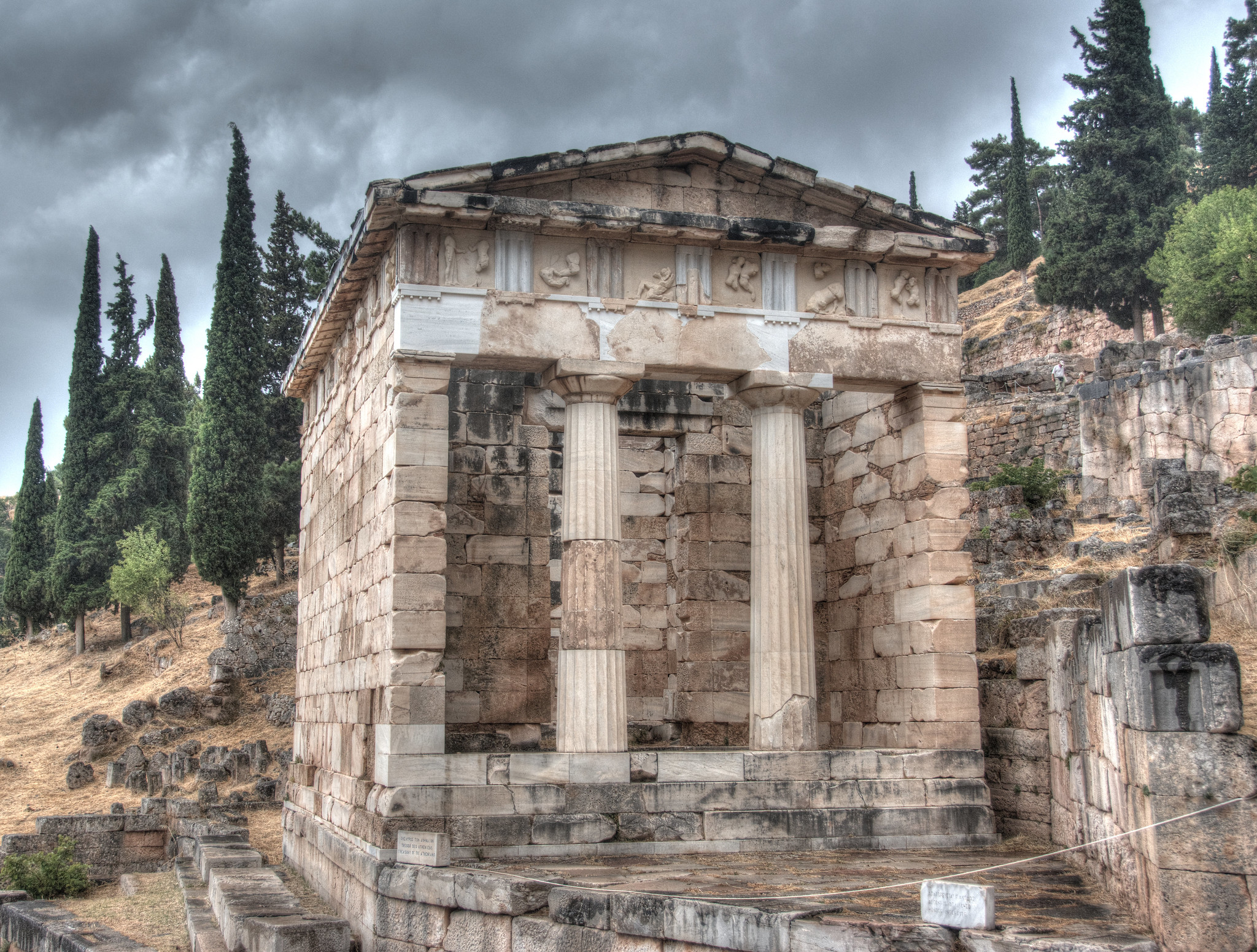 Archaeological remains of the Athenian treasury on the hillside at Delphi. It is a small square buildings with walls on three sides, and open with two columns at the front. The columns are partially reconstructed. The metopes are decorated with reliefs of figures.