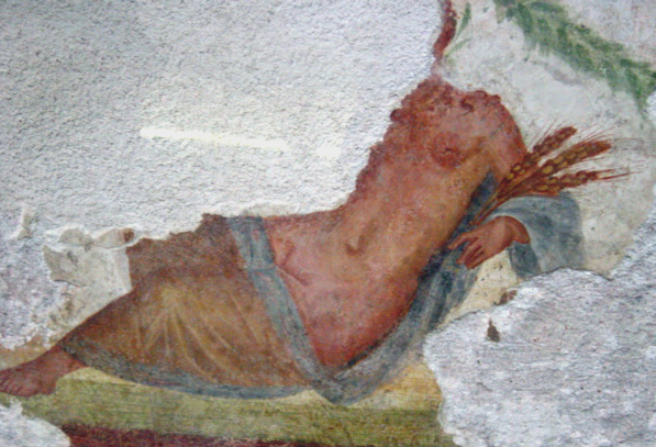 The Goddess Ceres reclines on a bright green couch, frontal to the viewer. Her head and right arm have chipped off. The rest of her body from the neck down is visible. She is nude to her upper thighs, which are covered with a translucent, blue draped garment. Her left foot peeks out from the garment. She holds three sheaves of wheat in her left hand. Her skin is mid-brown, reddish, her breasts are small with large areolas, her pubic area is undefined. The background is bright white. An evergreen garland crosses the upper right corner of the scene.