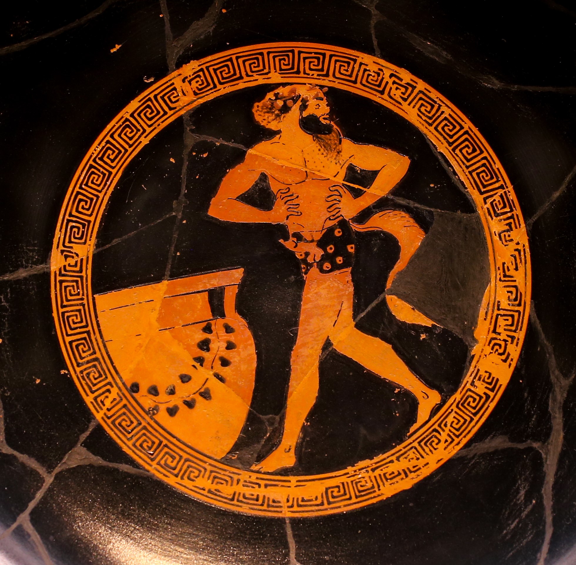 A bearded actor dressed as an ithyphallic satyr, with a tail attached to his waist by a belt. He is walking with his hands on his hips and head turned to look behind him. A large jar stands on the ground in front of him.