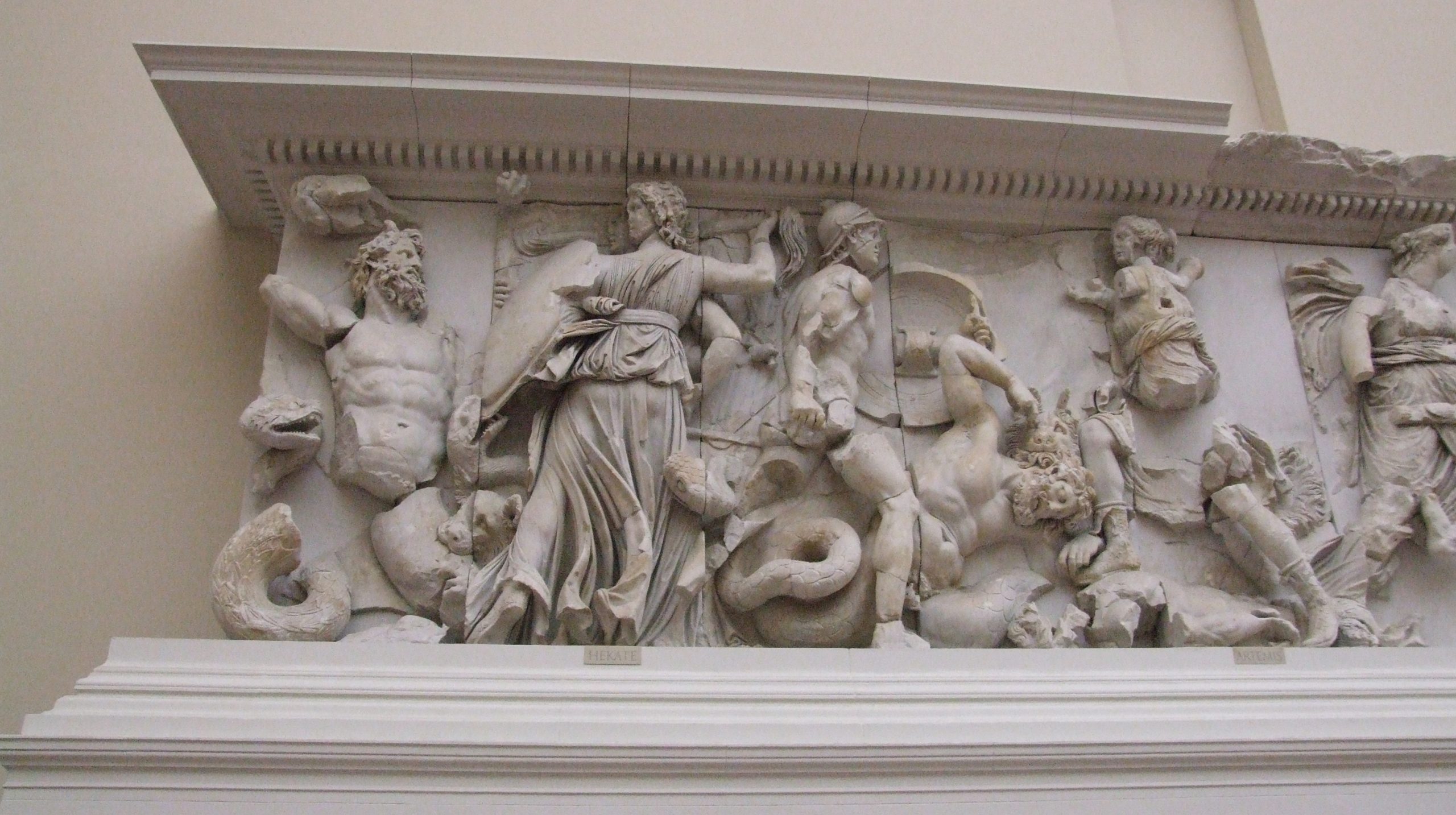 White marble frieze from the Altar of Pergamum. Left hand scene shows Hecate on the right with shield and brandishing torch. To her left the monster Clytios, with snakes for legs is attacking. One of his snake legs bites Hecate's shield. One of Hecate's dogs bites at the lower torso of Clytias.
