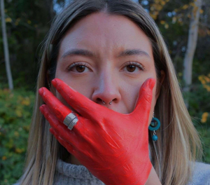 Image of a woman with her hand painted red and placed over her mouth.