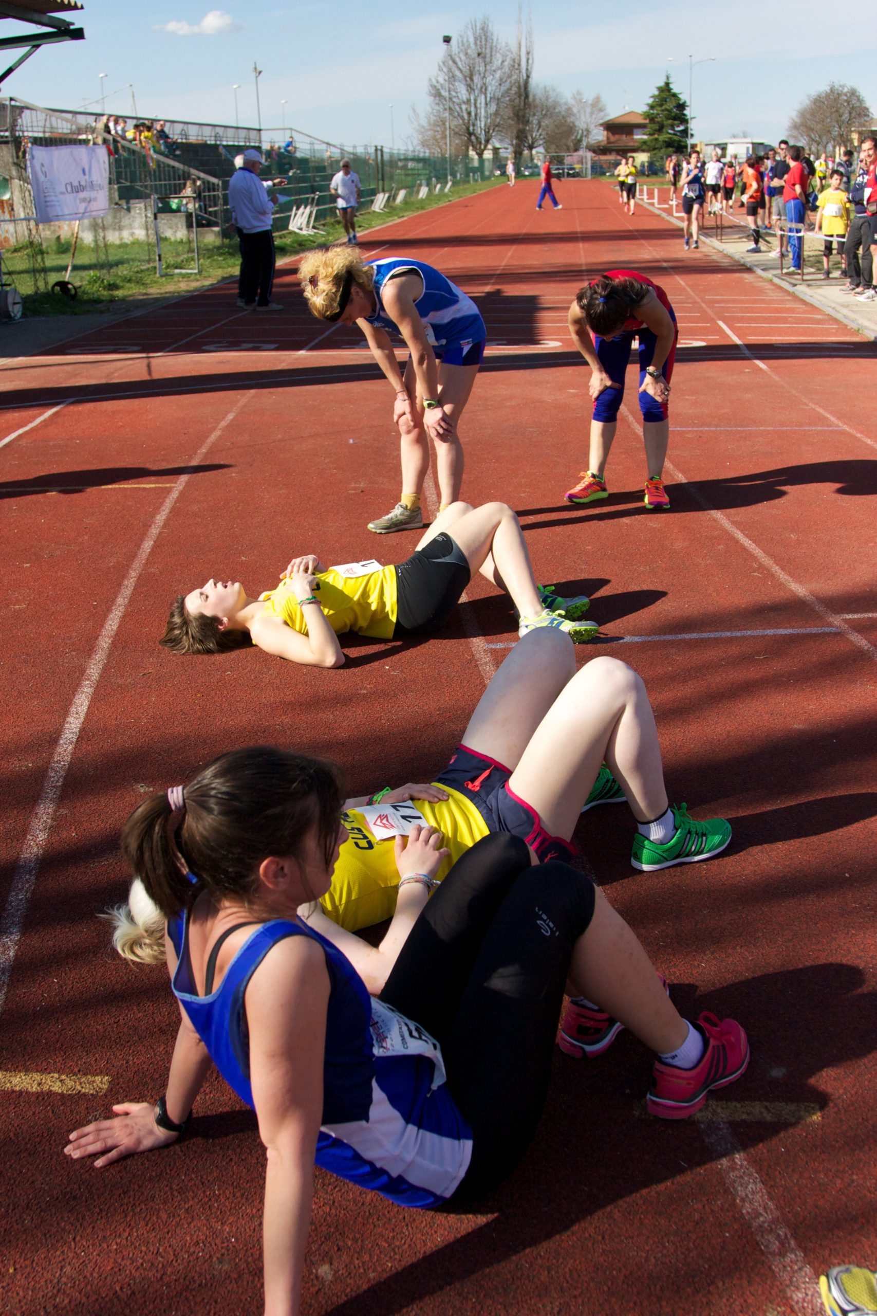 Image shows female track and field runners resting after a race. Three women are resting on the ground and two are leaning over with their hands on their knees, catching their breathe.