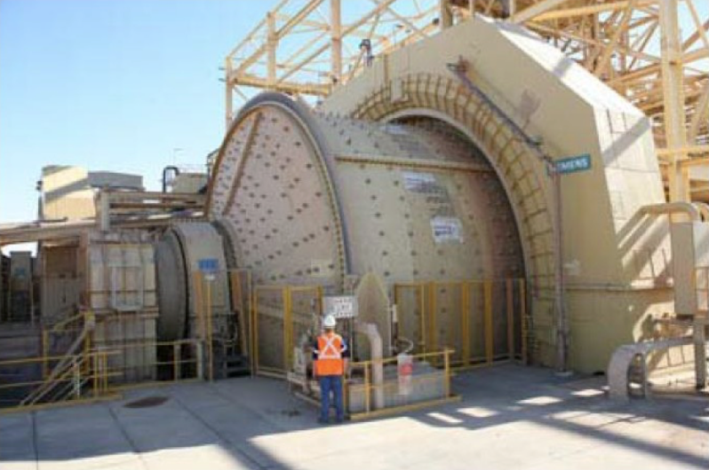 Photograph of a large semi‑autogenous grinding (SAG) mill in a mining facility: massive horizontal cylindrical drum with Siemens label, surrounded by safety railings. A worker in high‑visibility vest and helmet stands in front, showing the scale of the equipment.
