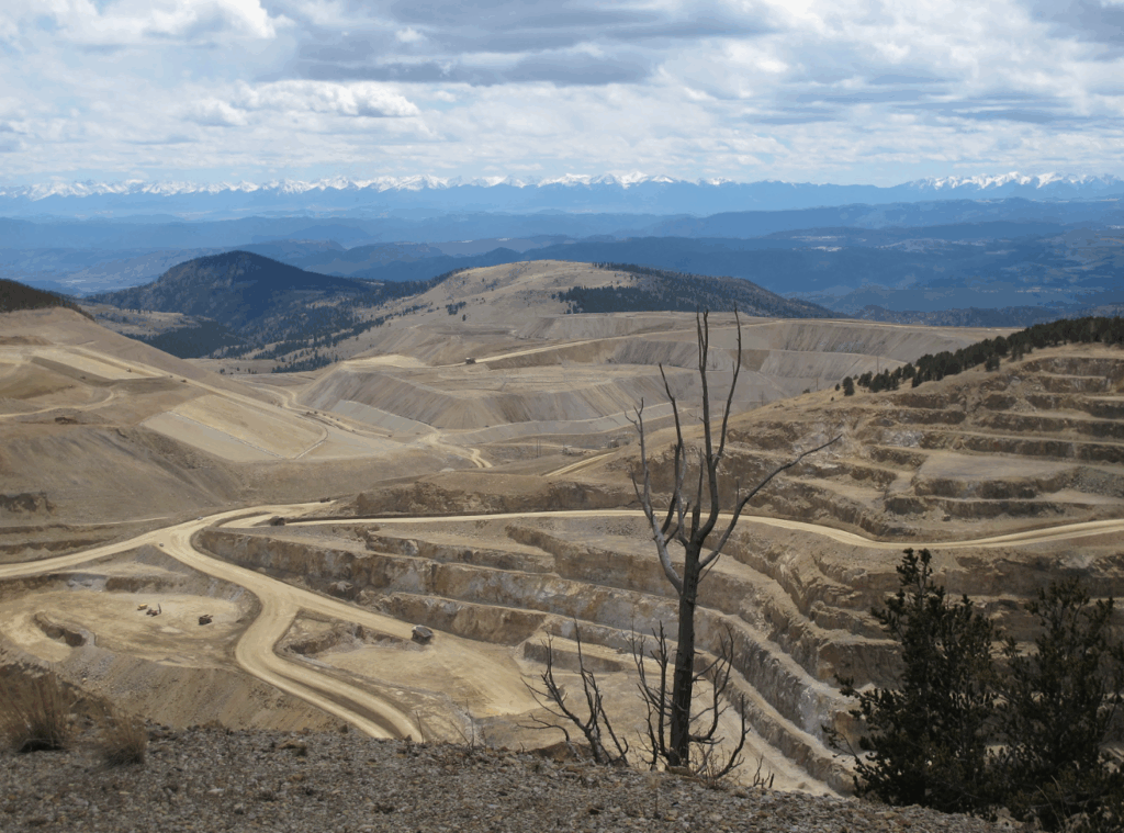 Large open‑pit mine in a mountainous areaof Cresson Pit, with terraced benches and spiral roads descending into the excavation. Foreground shows sparse vegetation and exposed rock; background features forested hills and distant snow‑capped peaks under a partly cloudy sky, highlighting the scale and environmental impact of the mining operation.