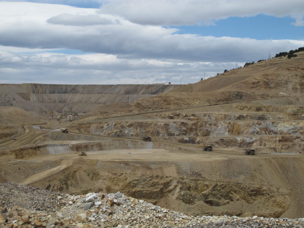 Large open‑pit mine with multiple terraced levels and winding haul roads. Several massive mining trucks travel along the benches, emphasizing the scale of the operation.
