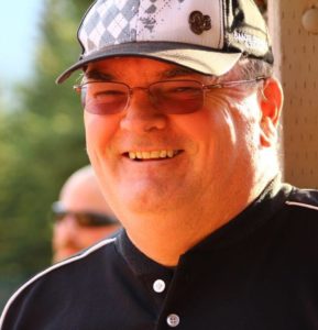 A man in a baseball cap and sunglasses smiles outside.