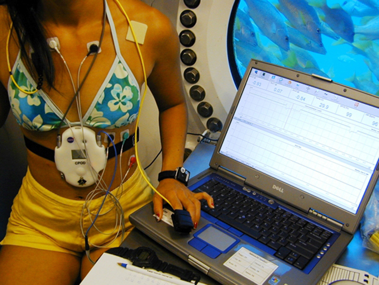 Photograph of a NASA scientist in an underwater habitat recording her vital signs using a portable device and a laptop computer.