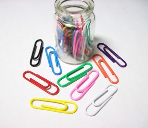 This picture shows a jar of coloured paperclips with several paperclips on the table around it.