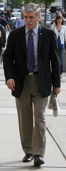 This picture shows Roméo Dallaire walking on the sidewalk on a busy city street.