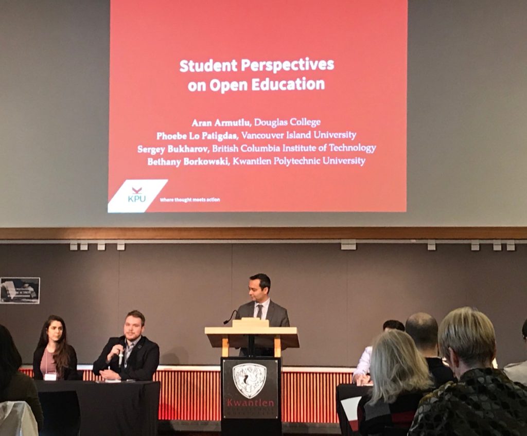 Four students seated with one moderator standing between them. One of the students is speaking. The screen above the group reads "Student Perspectives on Open Education"