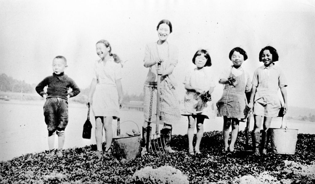 In this photo, Hanako Sato and a group of five smiling children at Vancouver Japanese Language School pose with a picnic basket and garden implements.