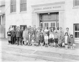 Shown is a photo of a group in front of Catholic Japanese Mission. Two nuns stand behind a group of about two dozen Japanese men, women, and children.