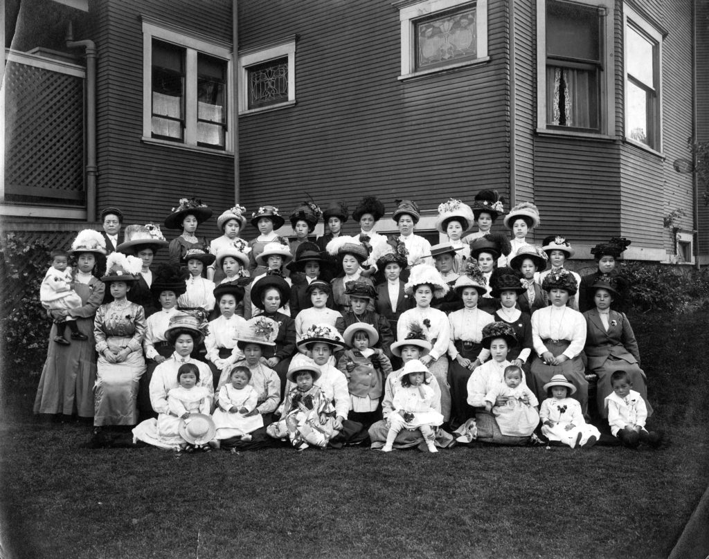 This is a group photo of the Japanese Women’s Association from around 1910. The women in the photo wear fine dresses and fancy hats with flowers, which suggests that prominent women took part in this association.