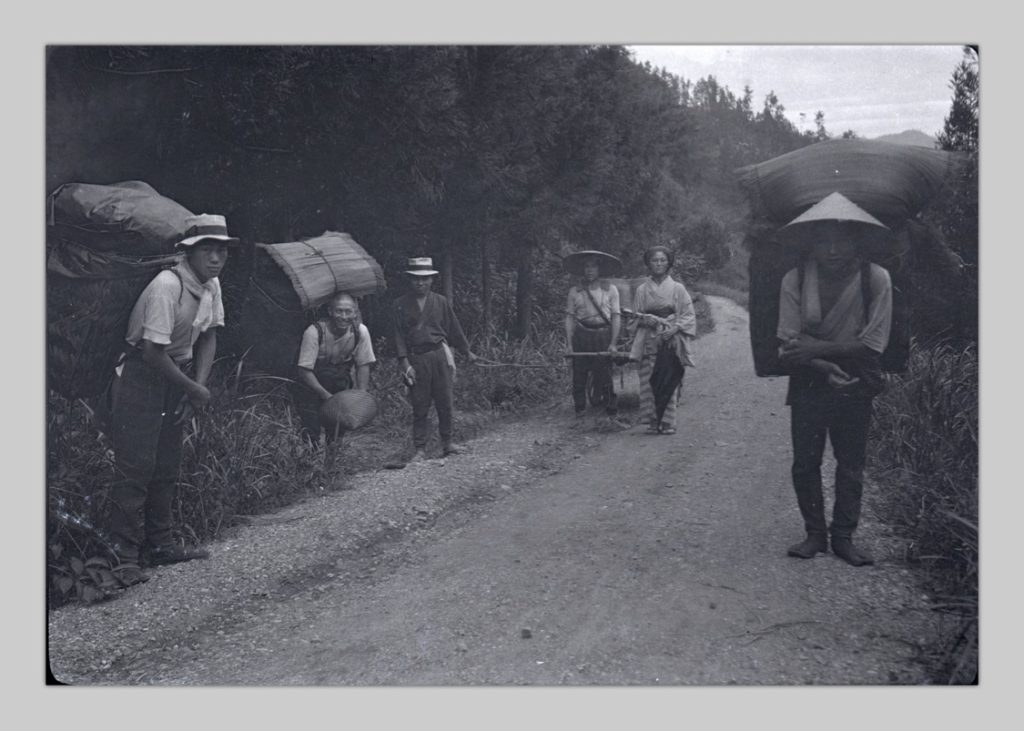 This is a photo, taken by John Cooper Robinson, of villagers carrying large rucksacks on their backs up a dirt-and-gravel road.