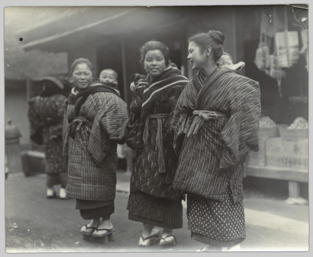 In this photo, three young women stand outside a market chatting. Their babies are swaddled in fabric, which is strapped securely to the mothers' outfits.