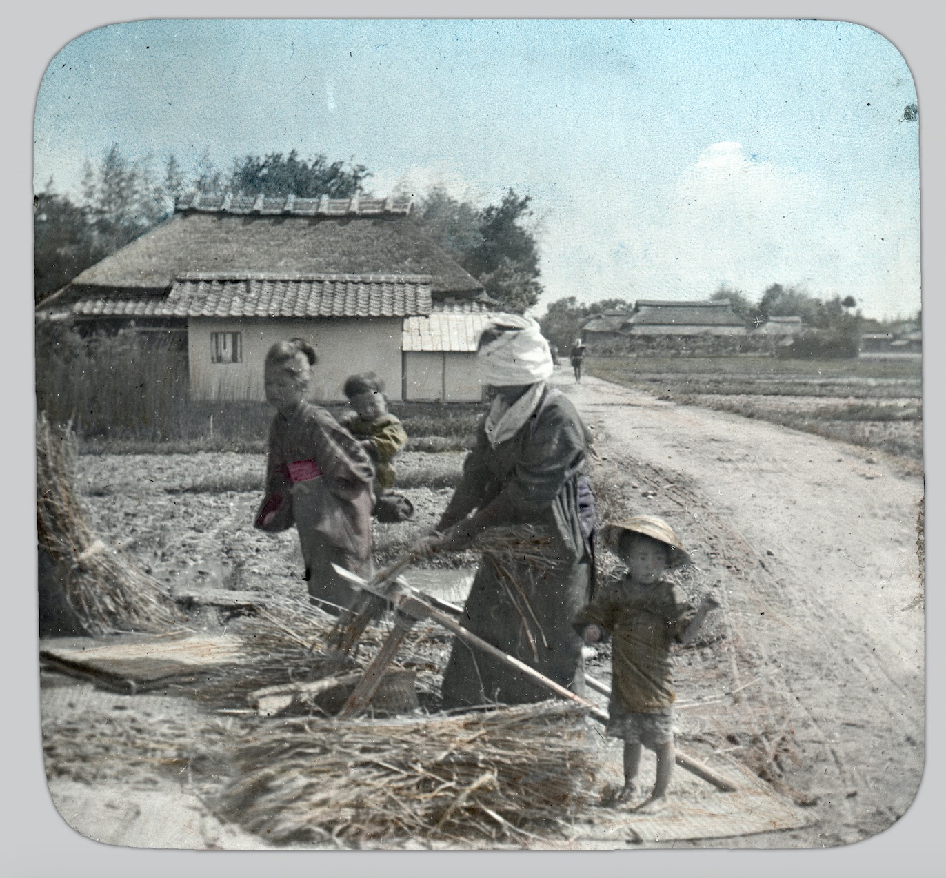 A colorized photo of two women collecting wheat. Two children also appear: one on one of the women's back and one standing next to a pile of cut wheat.