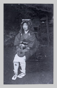 A glass negative copy of a commercial photograph of a Kyoto Oharame by John Cooper Robinson. A woman wearing sandals, kimono, and headdress sits upon a bundle of twigs.