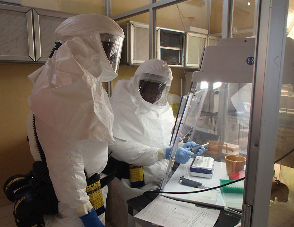 Two army medical officers in white hazmat suits sitting at a biosafety cabinet.
