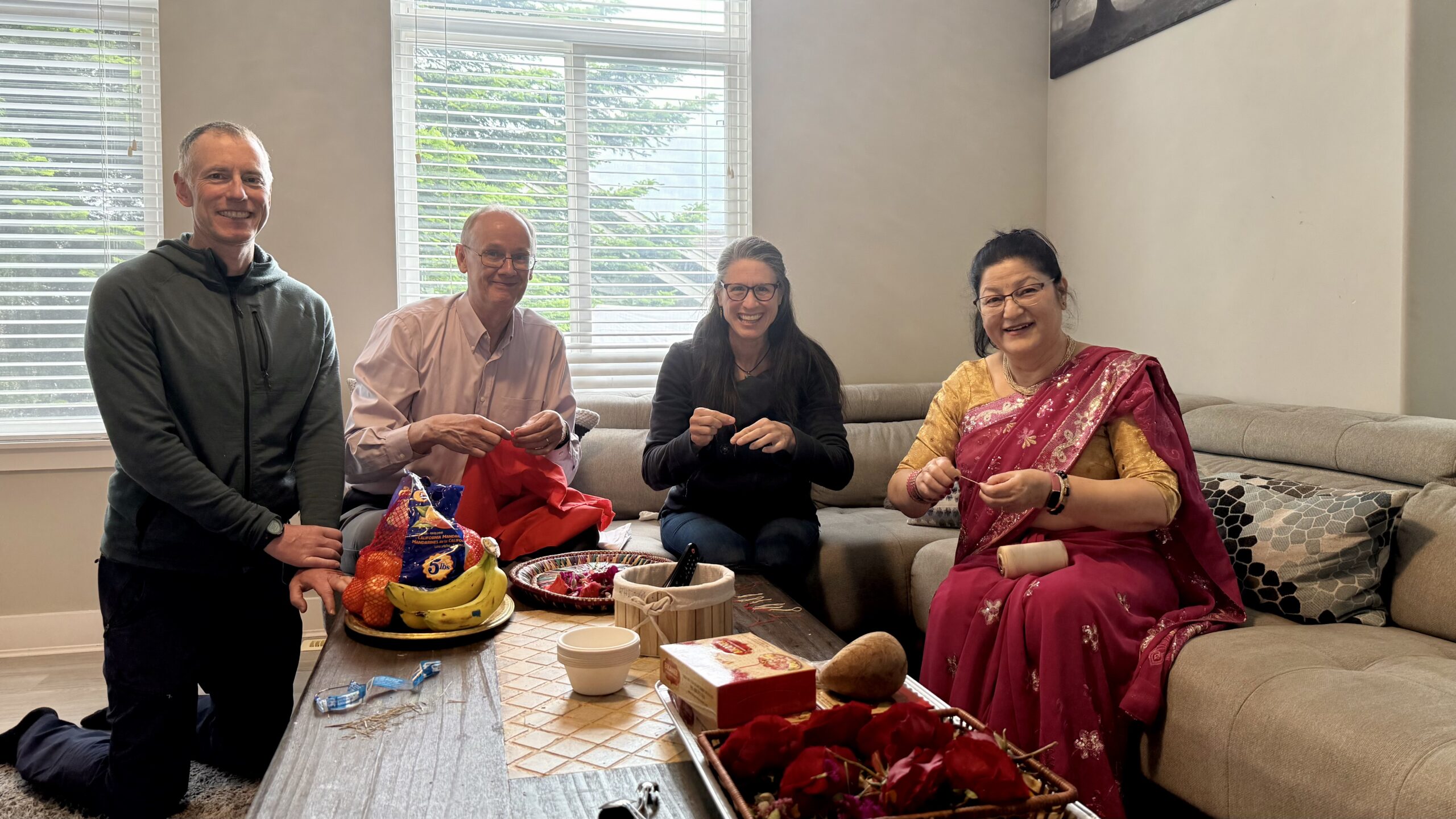 A group of four adults sit together in a living room, smiling and preparing small items at a coffee table.