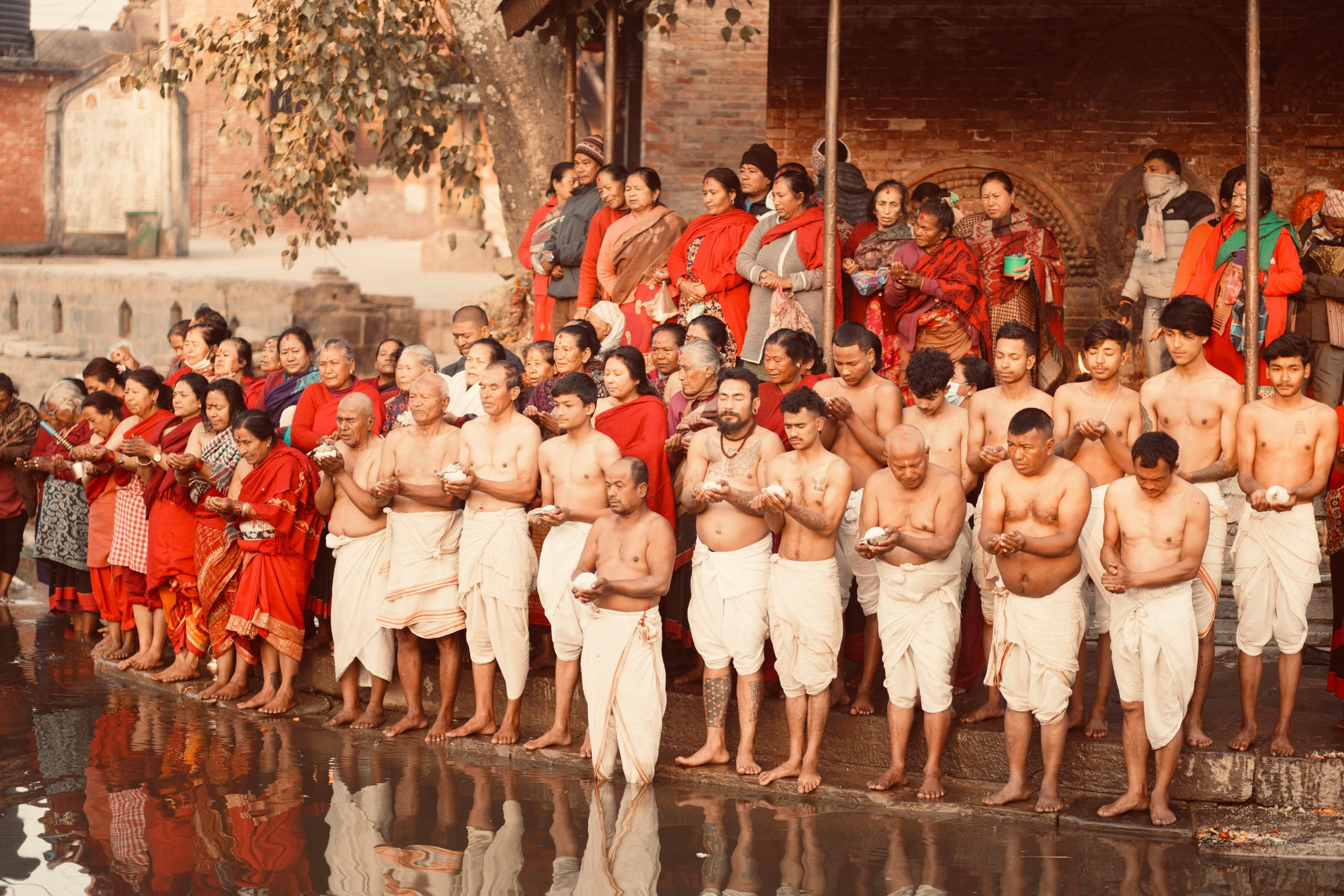 Devotees standing along a riverside in Bhaktapur, Nepal, performing a Hindu ritual; many wear traditional clothing.