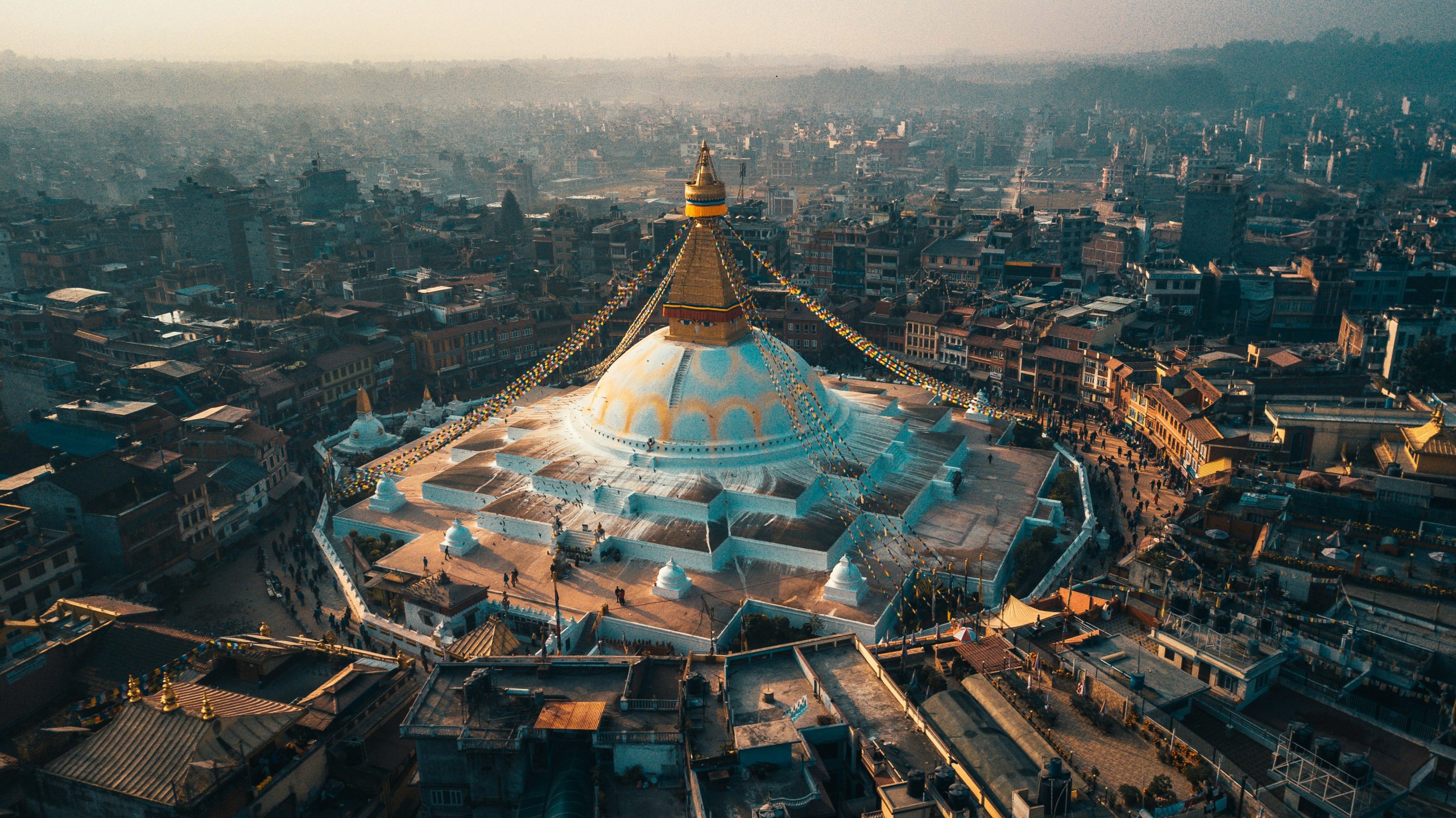 Aerial view of Boudhanath Stupa with prayer flags, surrounded by buildings in Kathmandu.