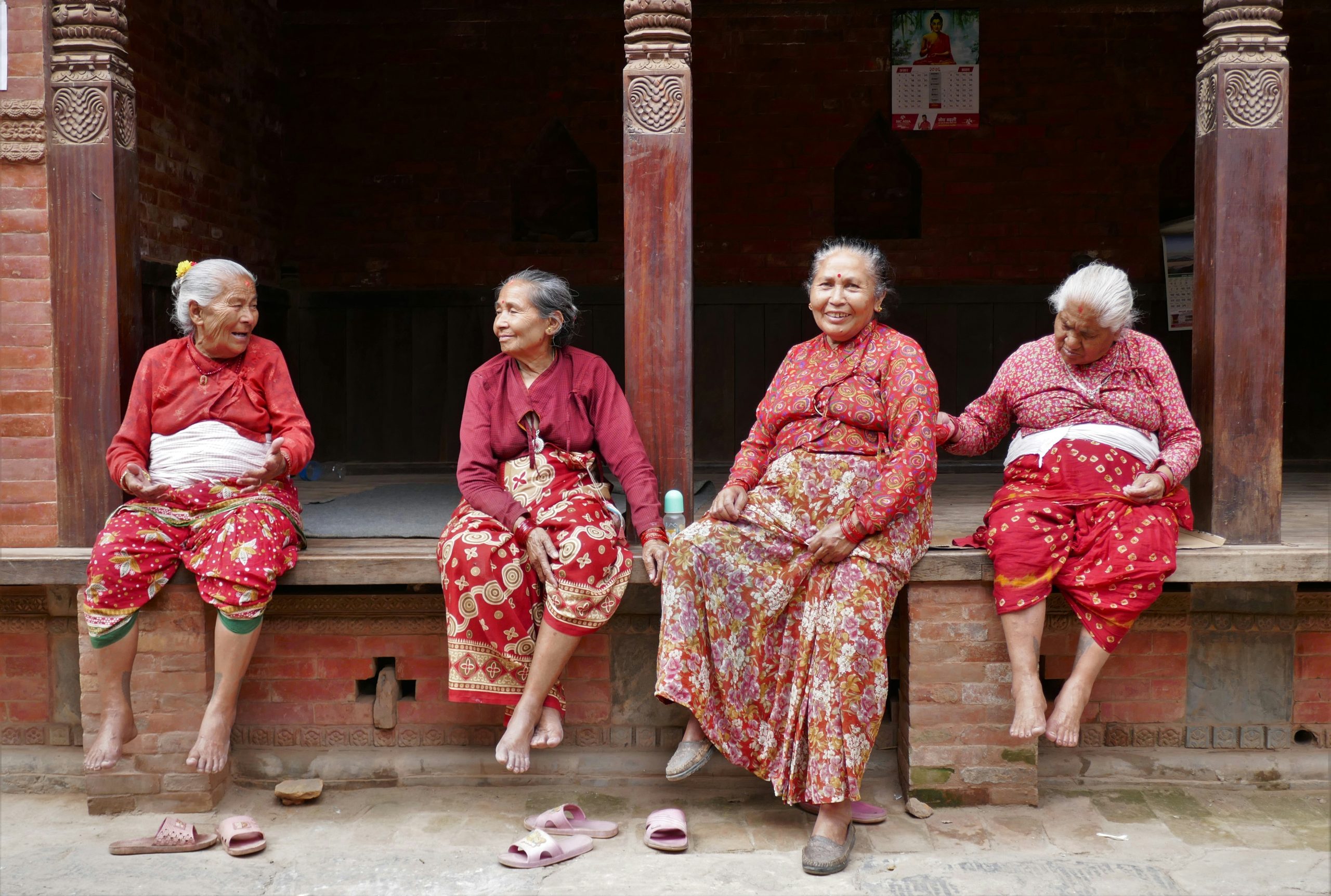 Four older women in colorful traditional clothing sit together on a raised wooden porch in a brick courtyard in Bhaktapur, Nepal.