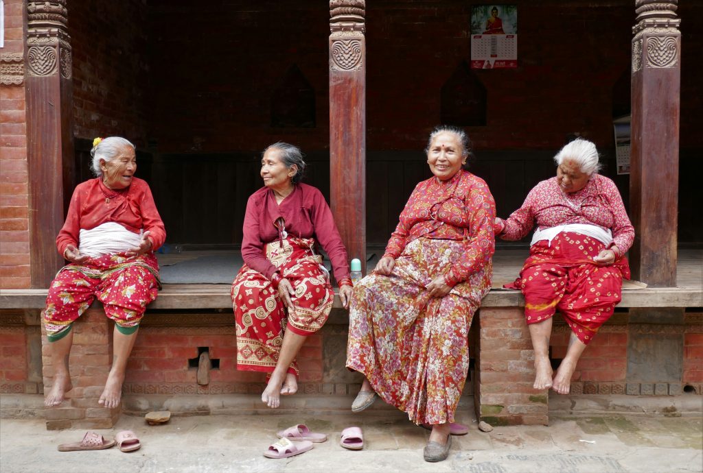 Four older women in colorful traditional clothing sit together on a raised wooden porch in a brick courtyard in Bhaktapur, Nepal.