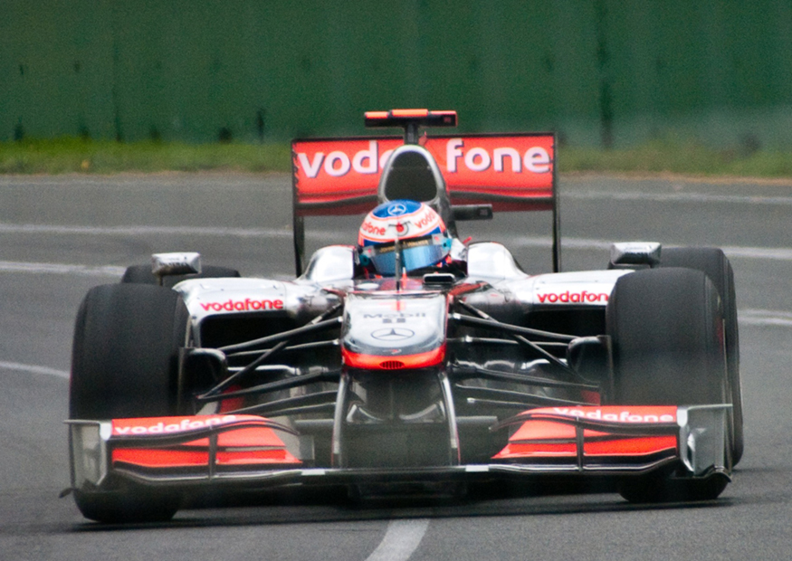 The figure shows, from front, a red and silver coloured Formula One car turning through a curve in a race on the Melbourne Grand Prix track, with the driver in seat.