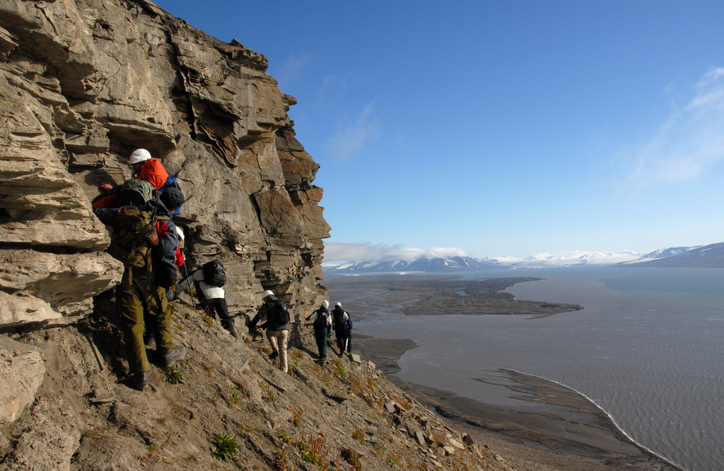 A line of people in hard hats walk along a steep slope at the base of a cliff. The landscape below opens to water.