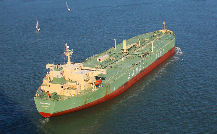 A photo of a supertanker in the water is shown. There are two much smaller vessels with sails in the distance.