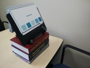 A desk with thee books stacked up and a book stand on top. On top of the book stand a spiral bound book is resting.